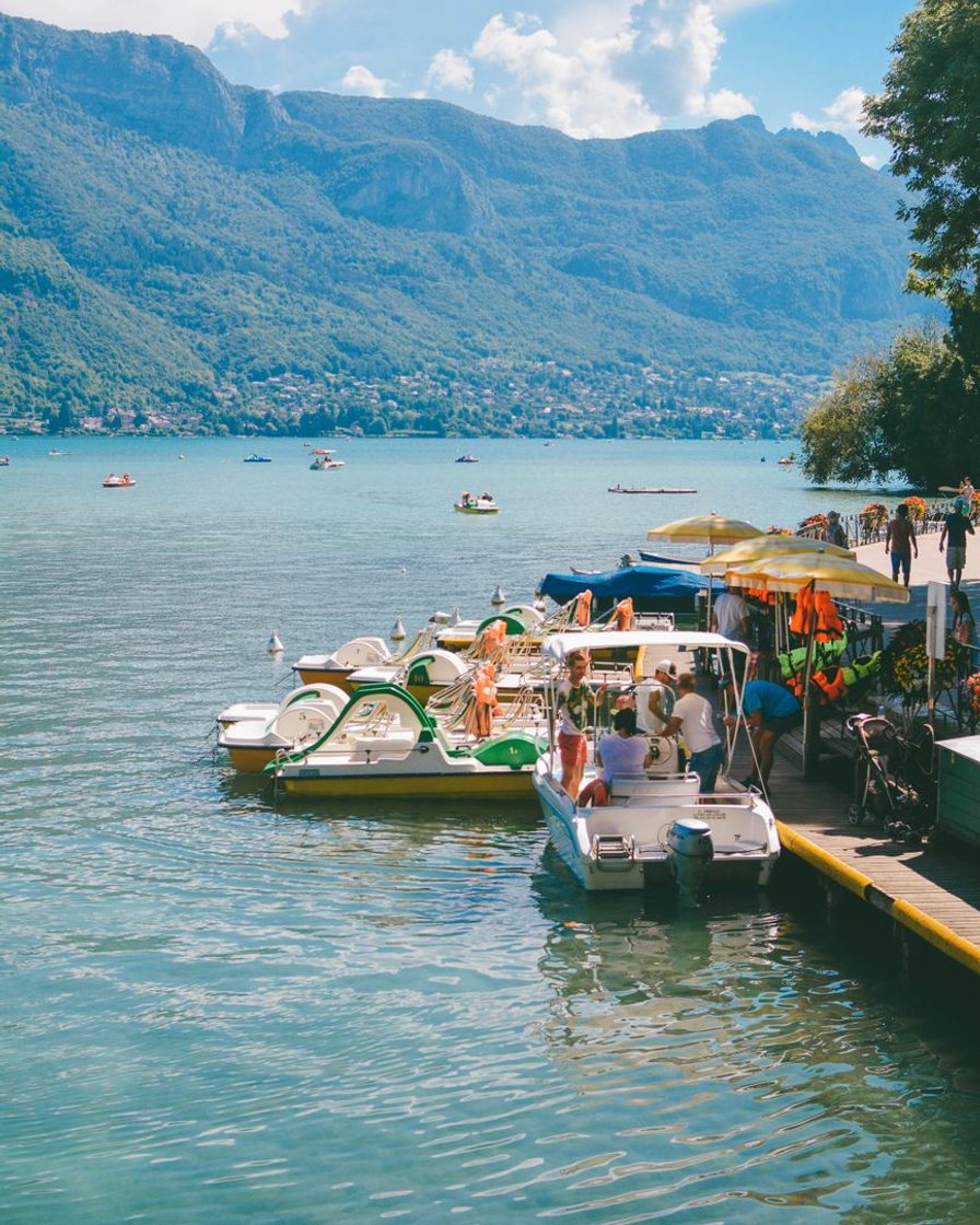Bateau de croisière blanc naviguant sur le lac d'Annecy devant les rives boisées de la rive est, passagers sur le pont supérieur, montagnes au fond