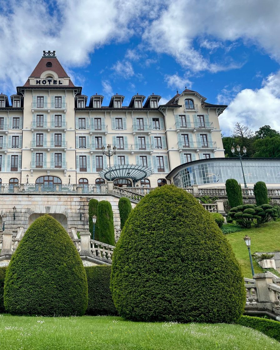 Façade d'un grand hôtel blanc face au lac d'Annecy avec terrasse-restaurant au bord de l'eau, parasols, pelouse descendant jusqu'à la rive, Semnoz en arrière-plan